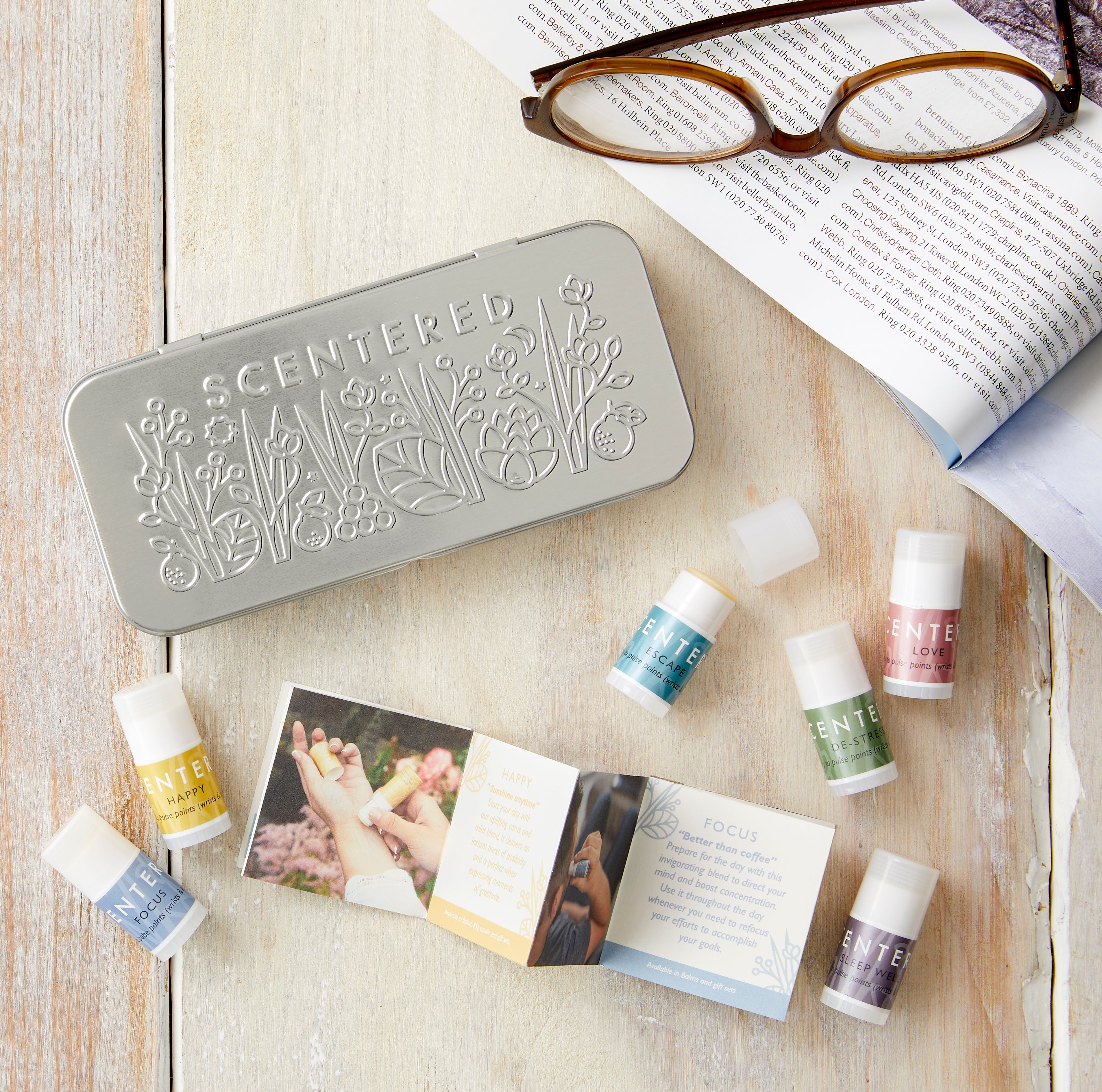 Scentered Discovery Tin featuring embossed floral pattern on the lid of the tin.  Next to the tin on a wooden table is a pair of glasses on an open magazine in the tone of relaxation.  There are also 6 Scentered aromatherapy balms shown scattered on the table which are the contents: Focus, Happy, Escape, De-stress, Love and Sleep Well Aromatherapy blends in 1.5g pulse point balm sticks