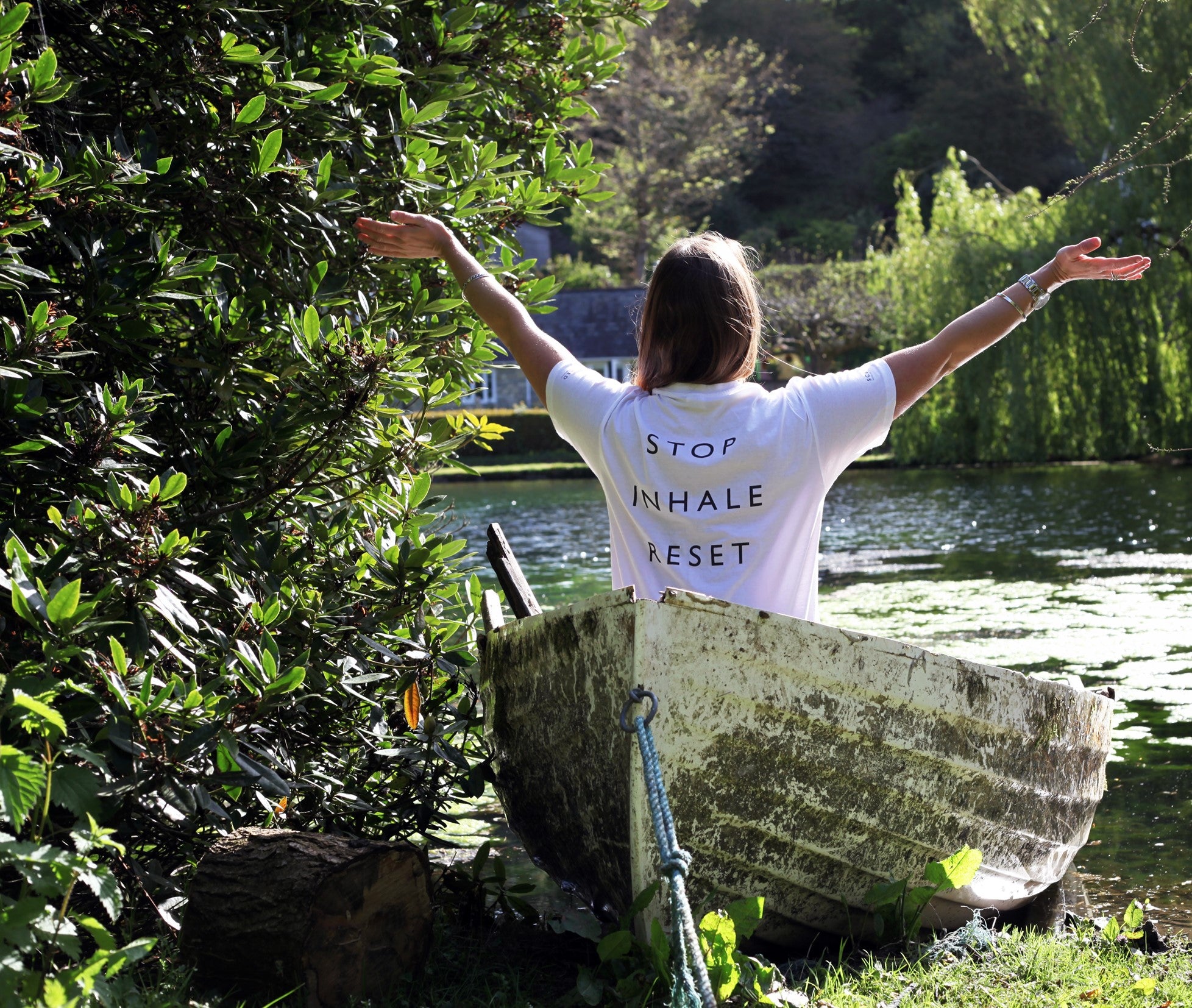 A women sitting in a boat on the river. Her arms out to the side and wearing a t-shirt with Stop, Inhale, Reset written on it.